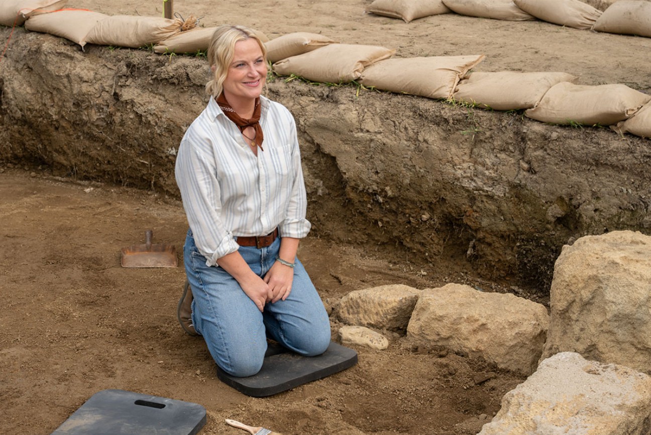 A woman kneels beside exposed stones at an archaeological excavation in Dig.