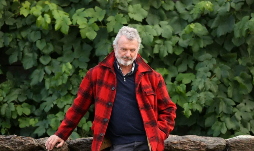 Sam Neill standing outdoors in a red plaid coat