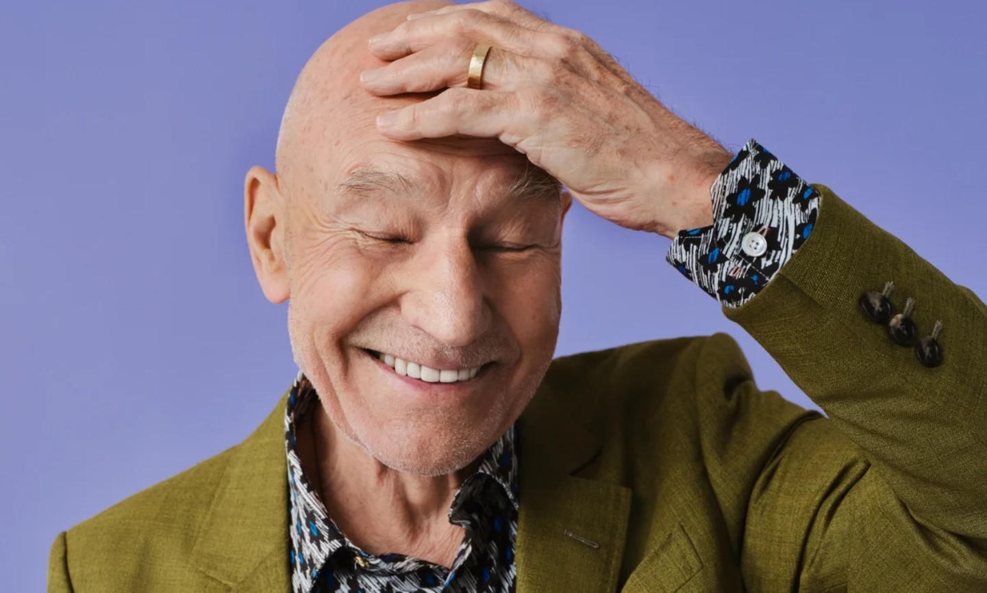 Patrick Stewart smiling with his eyes closed, one hand resting on his head, wearing an olive jacket against a lavender background